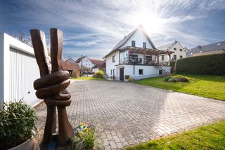 Frontalansicht auf ein Einfamilienhaus, im Vordergrund steht eine Holzskulptur.